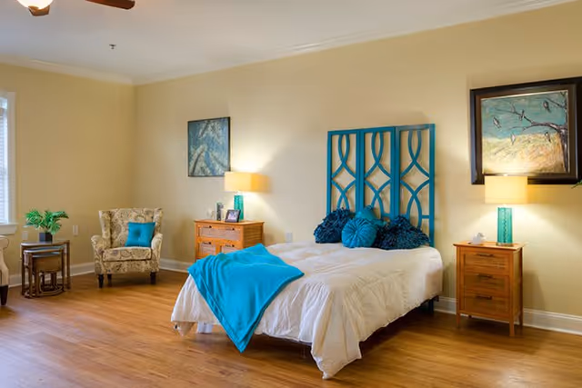 A bright and cozy bedroom with a single bed featuring a white comforter and blue decorative pillows and throw. The bed has a distinctive blue headboard. There are two wooden nightstands on either side of the bed, each with a lamp. A patterned armchair with a blue cushion is placed near a window with white blinds. The room has light yellow walls, wooden flooring, and two framed paintings on the walls.