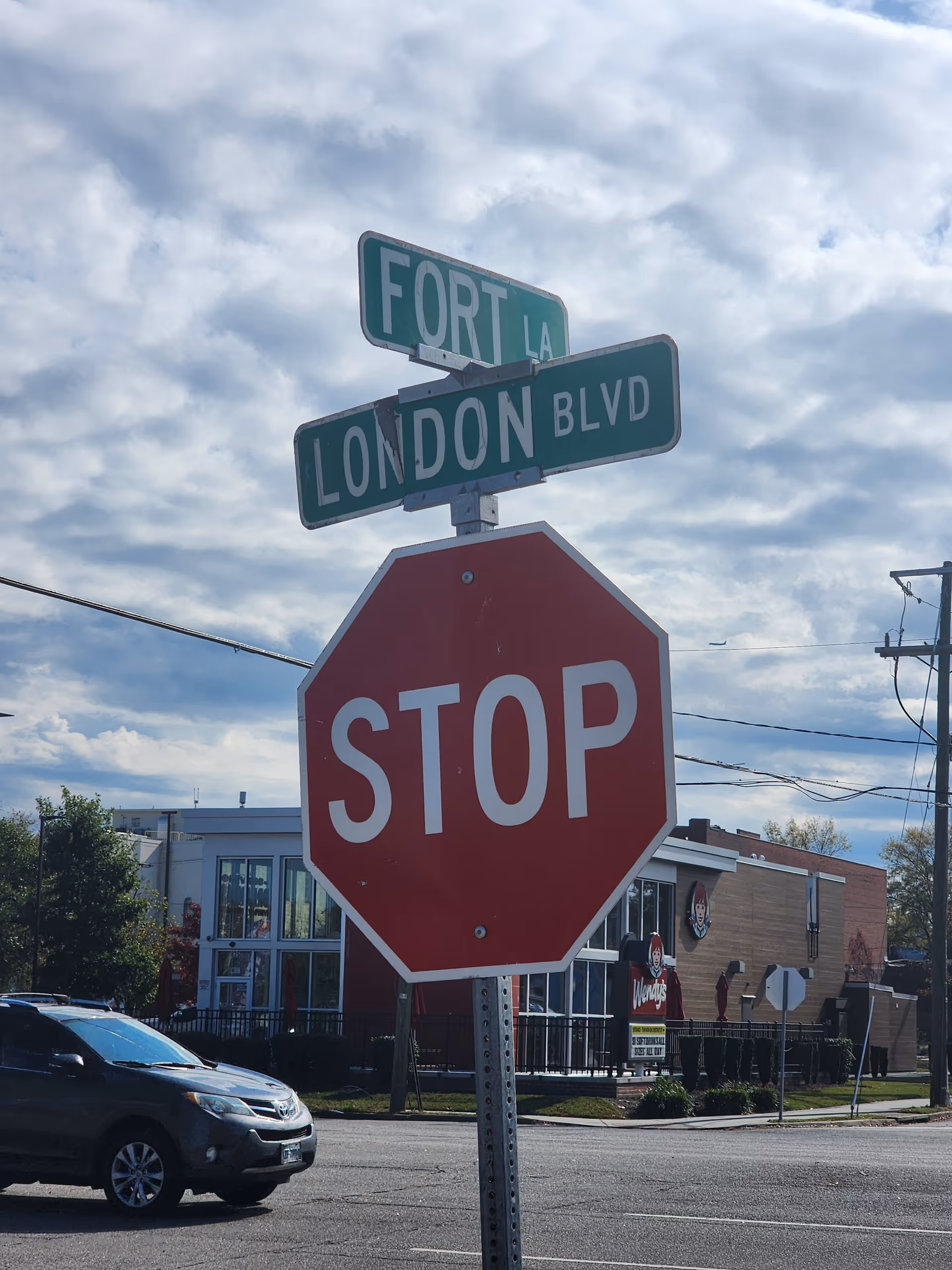 A stop sign at an intersection beneath green street signs reading 'LONDON BLVD' and 'FORT LA', with a Wendy's and other buildings in the background under a partly cloudy sky.