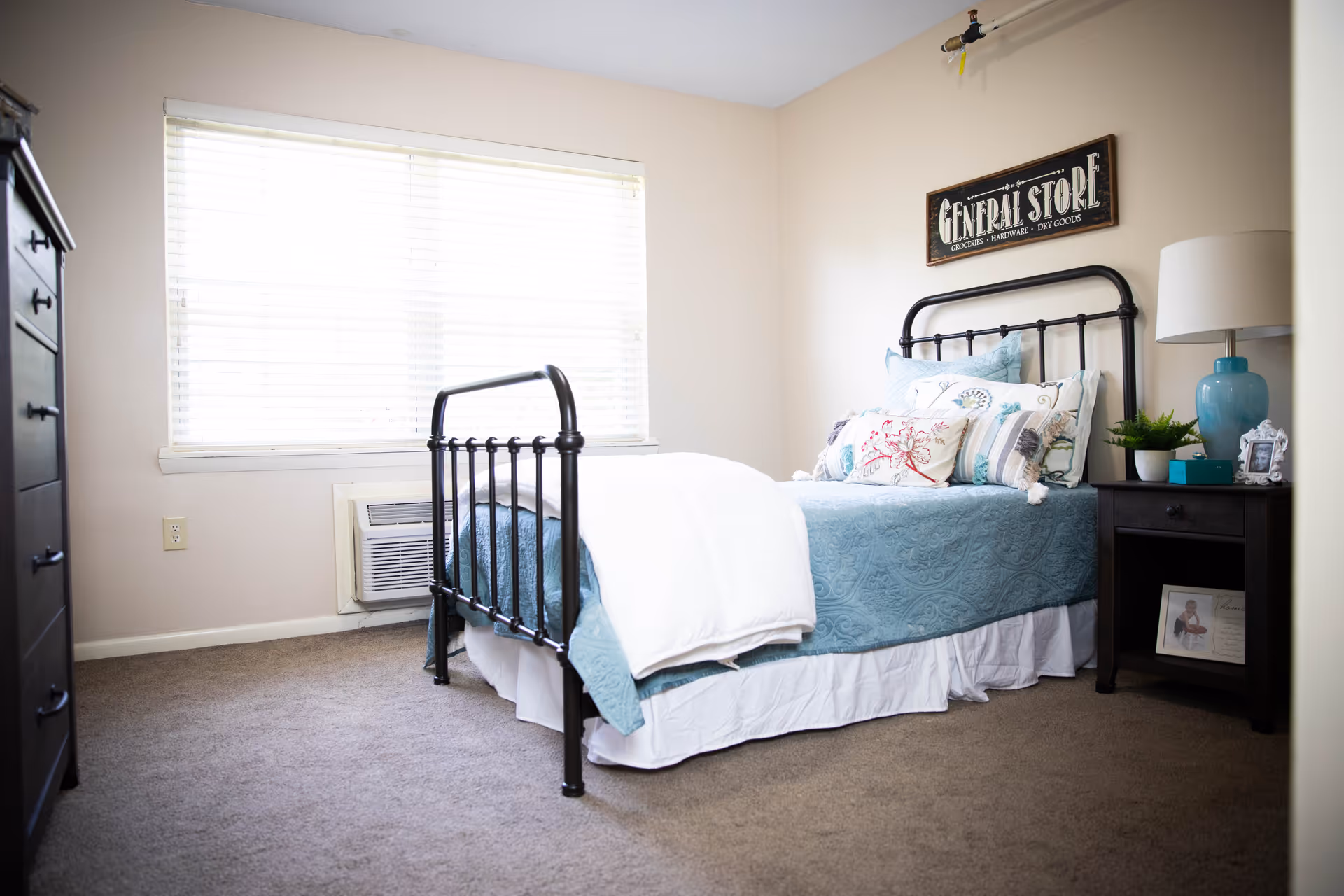 A cozy bedroom with a black metal bed frame, a bed made with a blue quilt and multiple decorative pillows. There is a nightstand with a blue lamp, a small plant, and a framed photo. A dresser is visible on the left side of the room. A large window with blinds lets in natural light. A wall sign above the bed reads 'GENERAL STORE'.