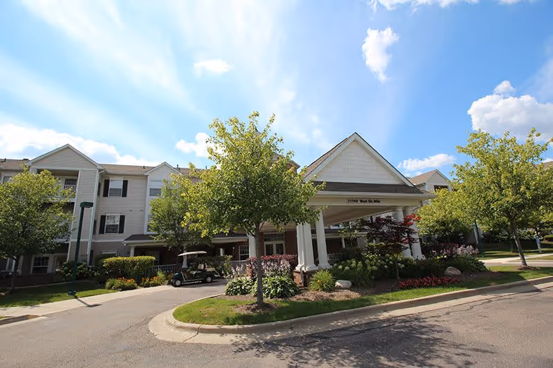 Front exterior of a three-story senior living building with a covered entrance, landscaped beds, and a parked golf cart.
