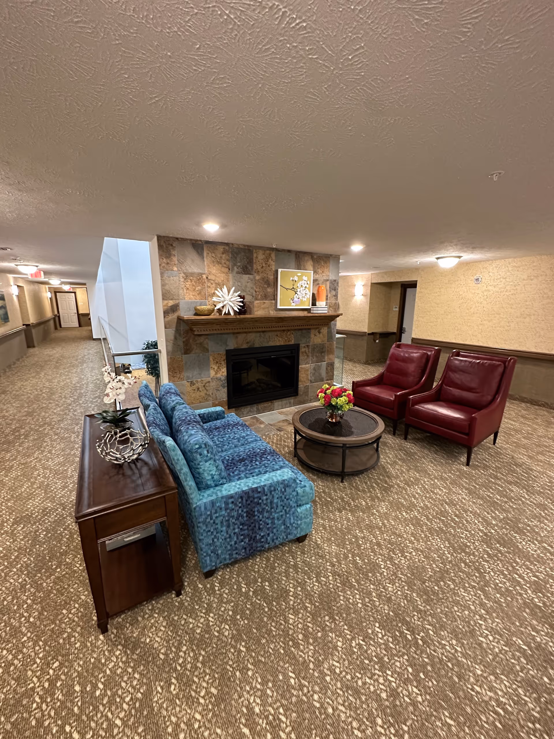 A seating area in a retirement community hallway with a blue sofa, two red armchairs, a round coffee table, and a tiled fireplace.