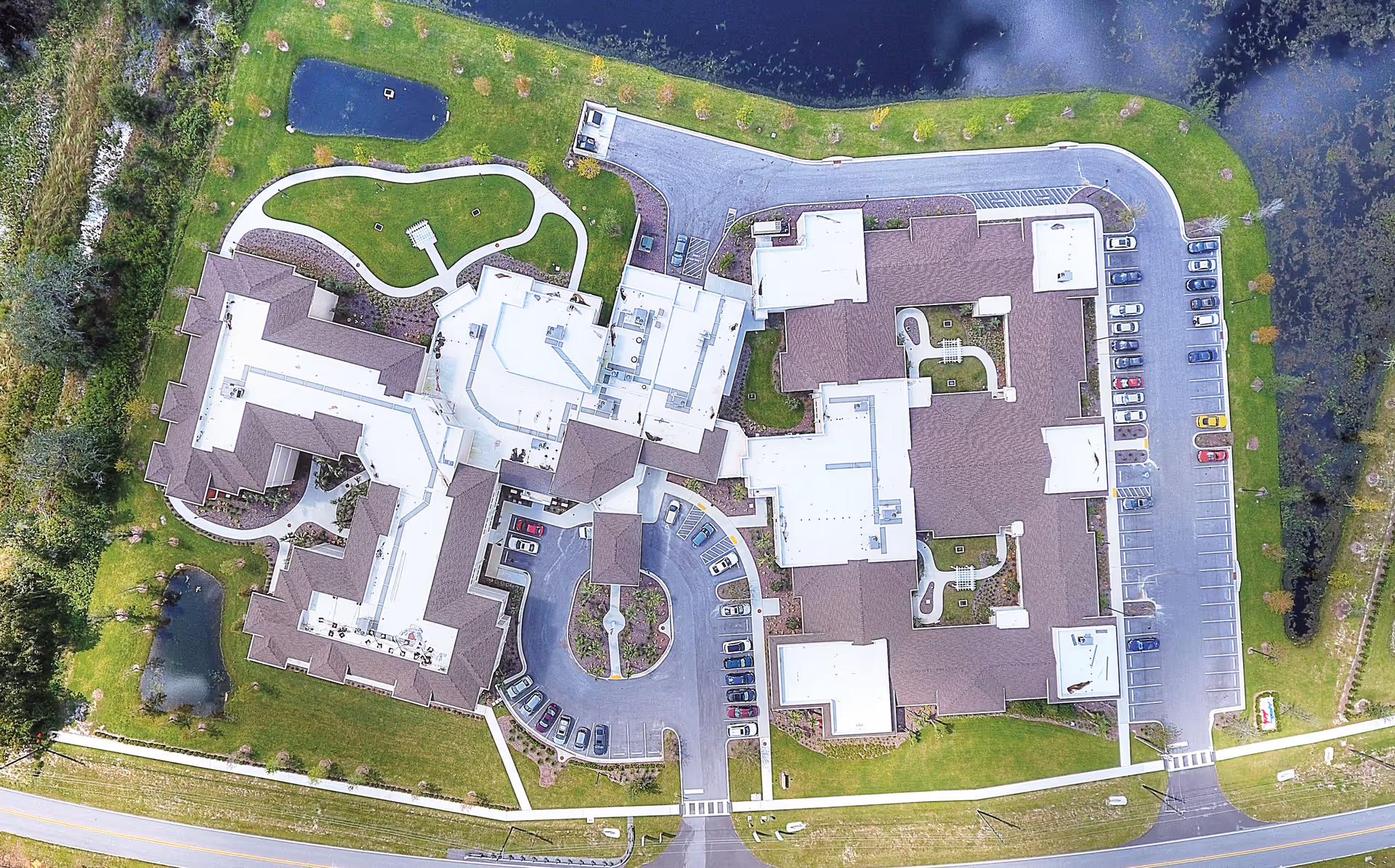 Aerial view of Promise Pointe Senior Living facility showing the building complex with multiple wings, surrounding parking lots, landscaped green areas, walking paths, and nearby water features.