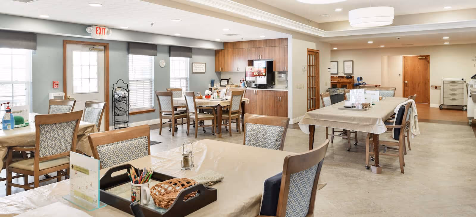 A bright and spacious dining room in a senior living facility with multiple tables and chairs arranged neatly. The room features large windows with blinds, a beverage station with a coffee machine, and various items like a basket, colored pencils, and a sanitizer bottle on the tables. The walls are painted light blue and white, and there is an exit door visible in the background.