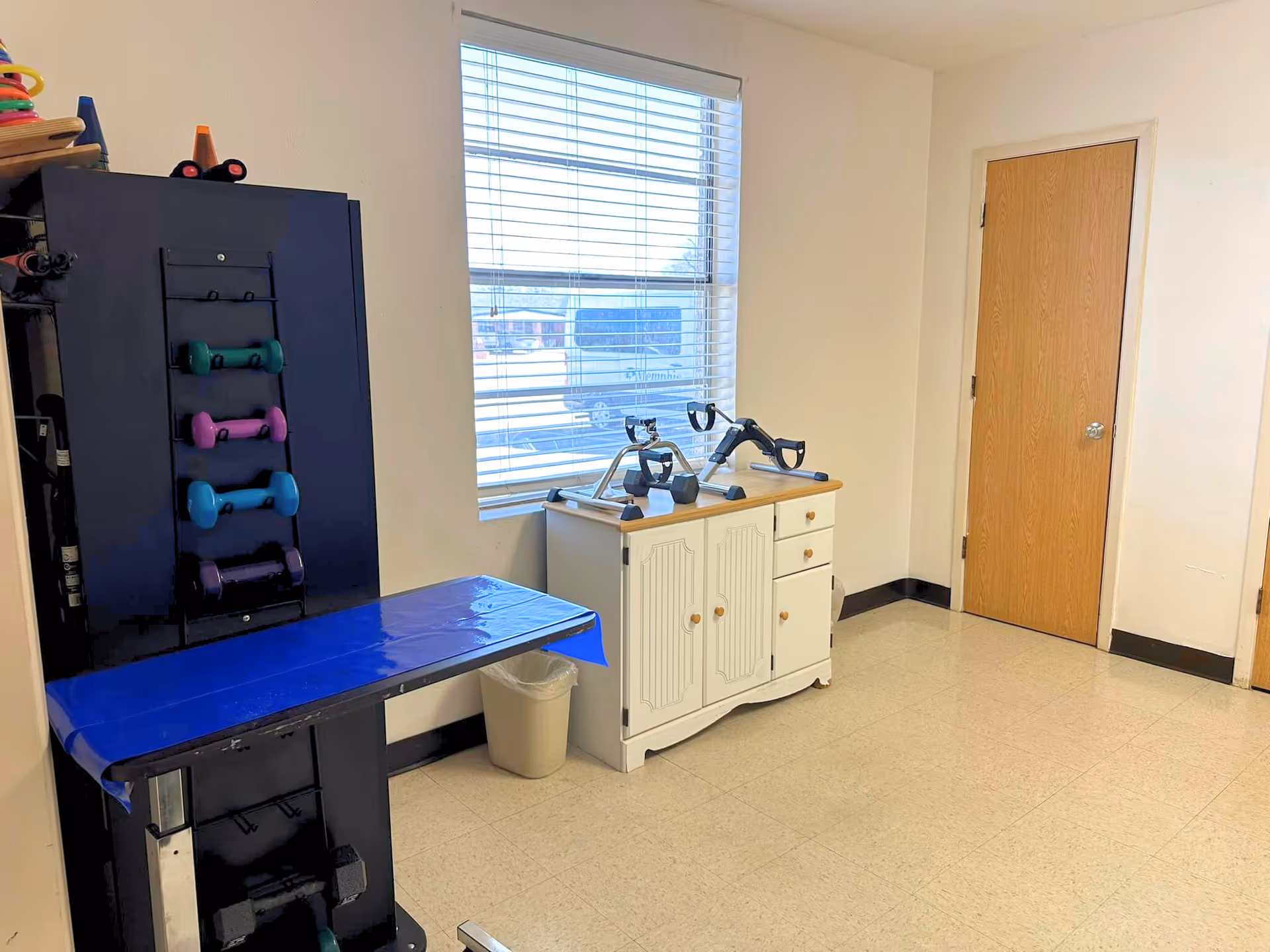 A small exercise room with a black exercise machine holding colorful dumbbells on the left, a white cabinet with exercise pedals on top under a window with blinds, and a closed wooden door on the right.