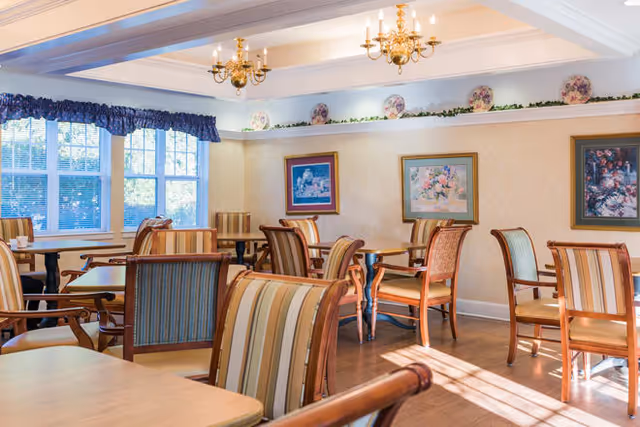 A bright dining room with multiple wooden tables and chairs featuring striped upholstery. The room has large windows with blue valances allowing natural light to fill the space. The walls are decorated with framed floral paintings and a decorative shelf with plates and greenery above.