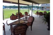 Covered outdoor patio with several tables and cushioned chairs overlooking a grassy lawn and nearby building.