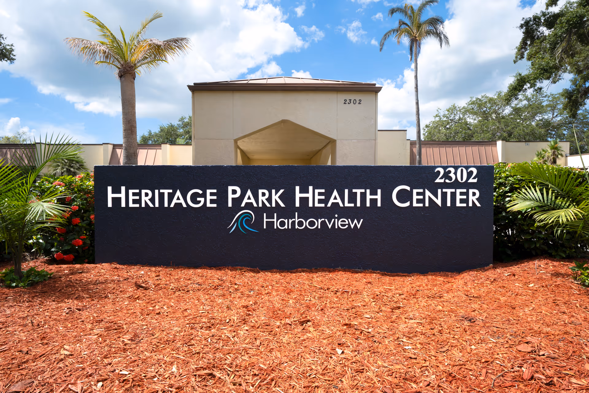 Exterior view of Heritage Park Health Center sign with the building entrance in the background, surrounded by palm trees and landscaping under a partly cloudy sky.