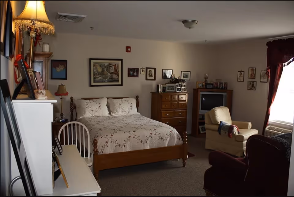A cozy bedroom in an assisted living facility featuring a wooden bed with floral bedding, a wooden dresser with framed photos, a TV on a stand, an armchair with a blanket, and a window with dark red curtains letting in natural light. The walls are decorated with various framed pictures and the room has a warm, inviting atmosphere.