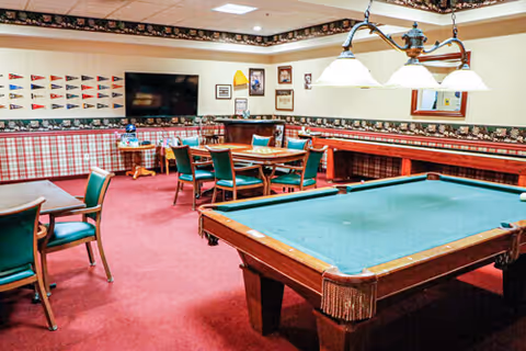 A recreational room with a green pool table in the foreground, several tables and chairs arranged around the room, a wall-mounted TV, and decorative flags and framed pictures on the walls. The room has red carpeting and patterned wallpaper borders near the ceiling.