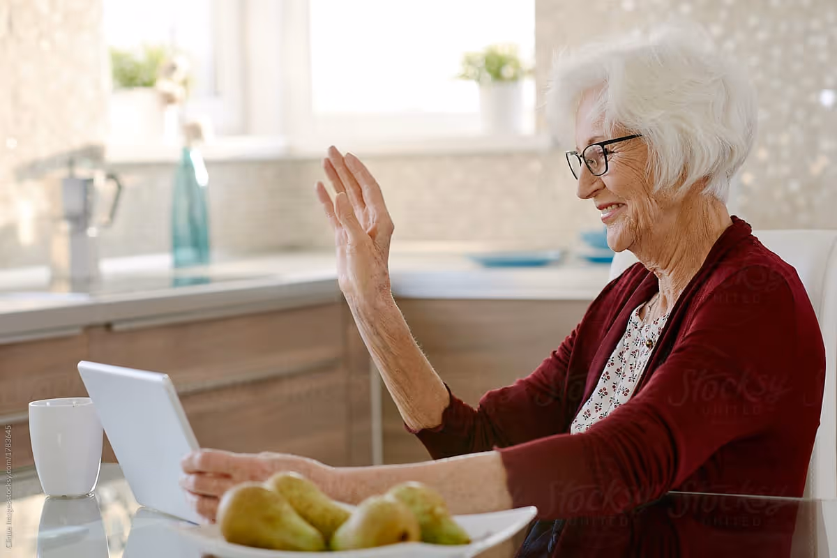 An elderly woman with white hair and glasses sitting at a kitchen table, smiling and waving at a tablet device. On the table in front of her are a white mug and a bowl of pears. The kitchen background is bright with natural light coming through the window.