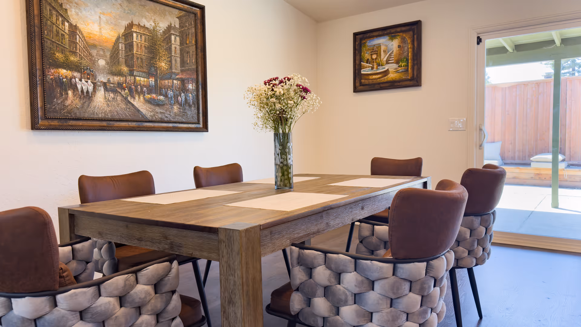 A dining room with a wooden table set with placemats and a vase of flowers in the center. Six cushioned chairs with a woven design surround the table. Two framed paintings hang on the walls, and a sliding glass door opens to an outdoor patio area with a wooden fence.