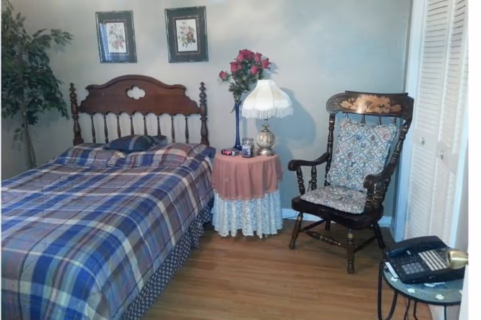 A bedroom with a wooden headboard bed covered in plaid bedding, a round side table with a lamp and flowers, and an ornate wooden chair.
