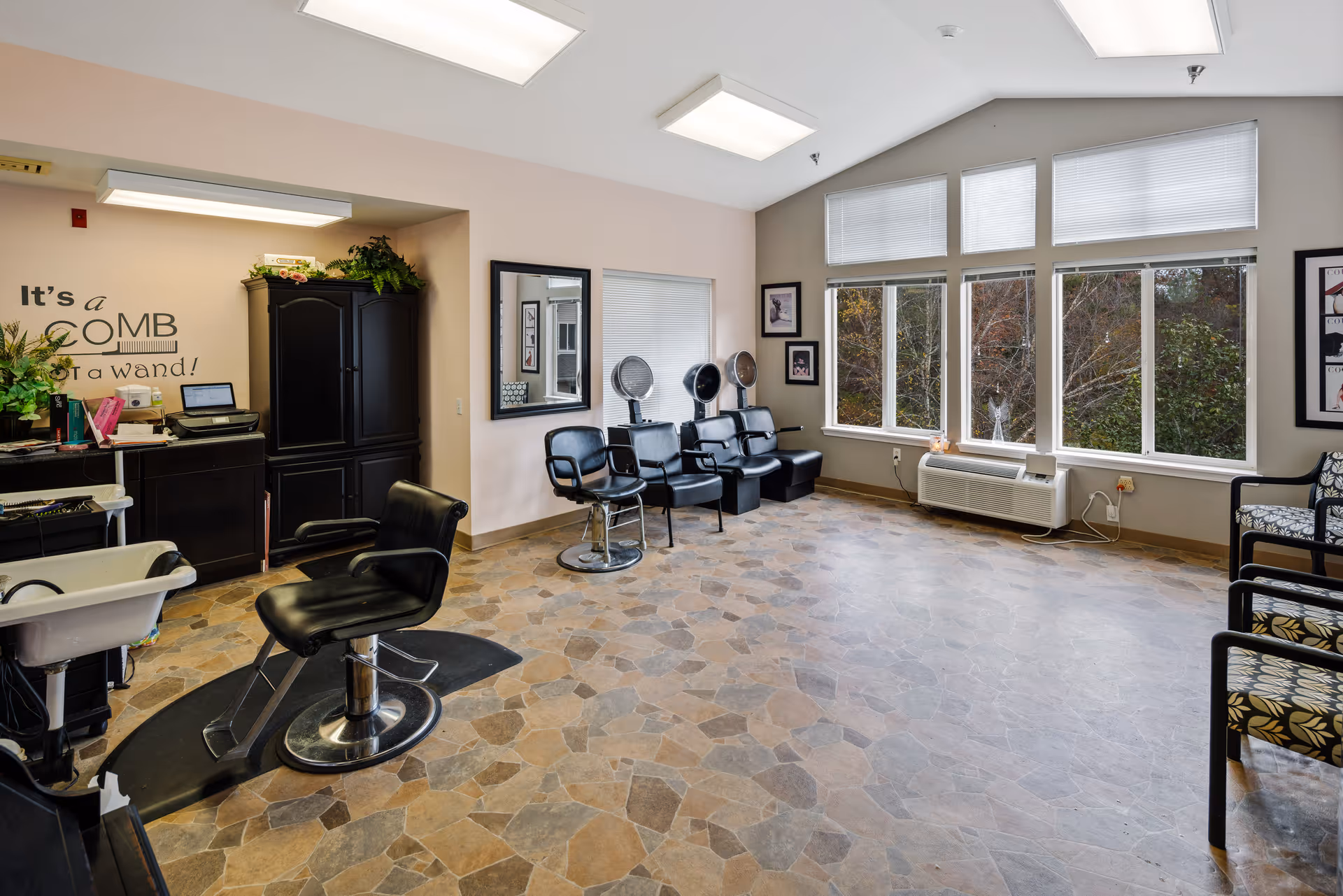 Interior of a senior living community hair salon with black salon chairs, hair dryers, a washing station, and large windows letting in natural light. The walls are light-colored, and there are framed pictures and plants decorating the space. A quote on the wall reads, 'It's a COMB not a wand!'