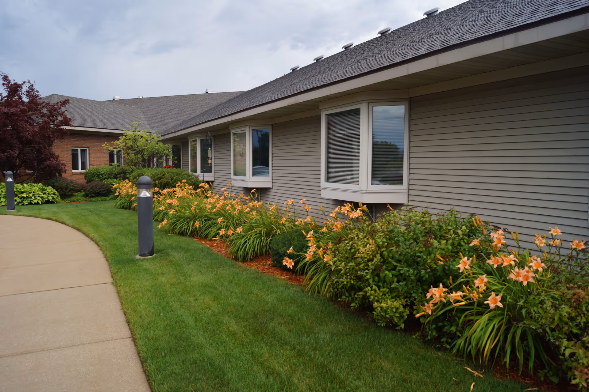 Single-story assisted living building exterior with a curved sidewalk, manicured lawn and beds of orange flowers.