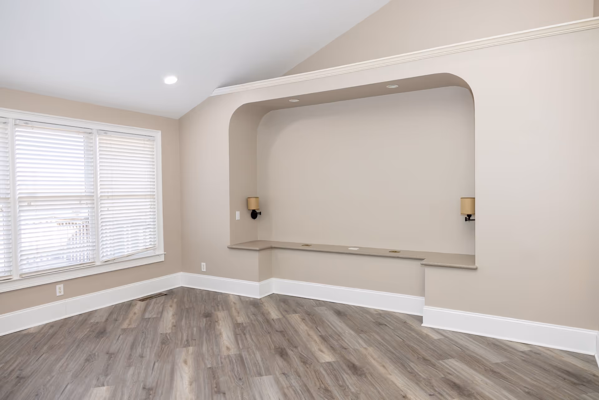Empty room with light beige walls, a large window with white blinds on the left, a recessed alcove with a built-in ledge and two wall-mounted lamps, and wood-look flooring.
