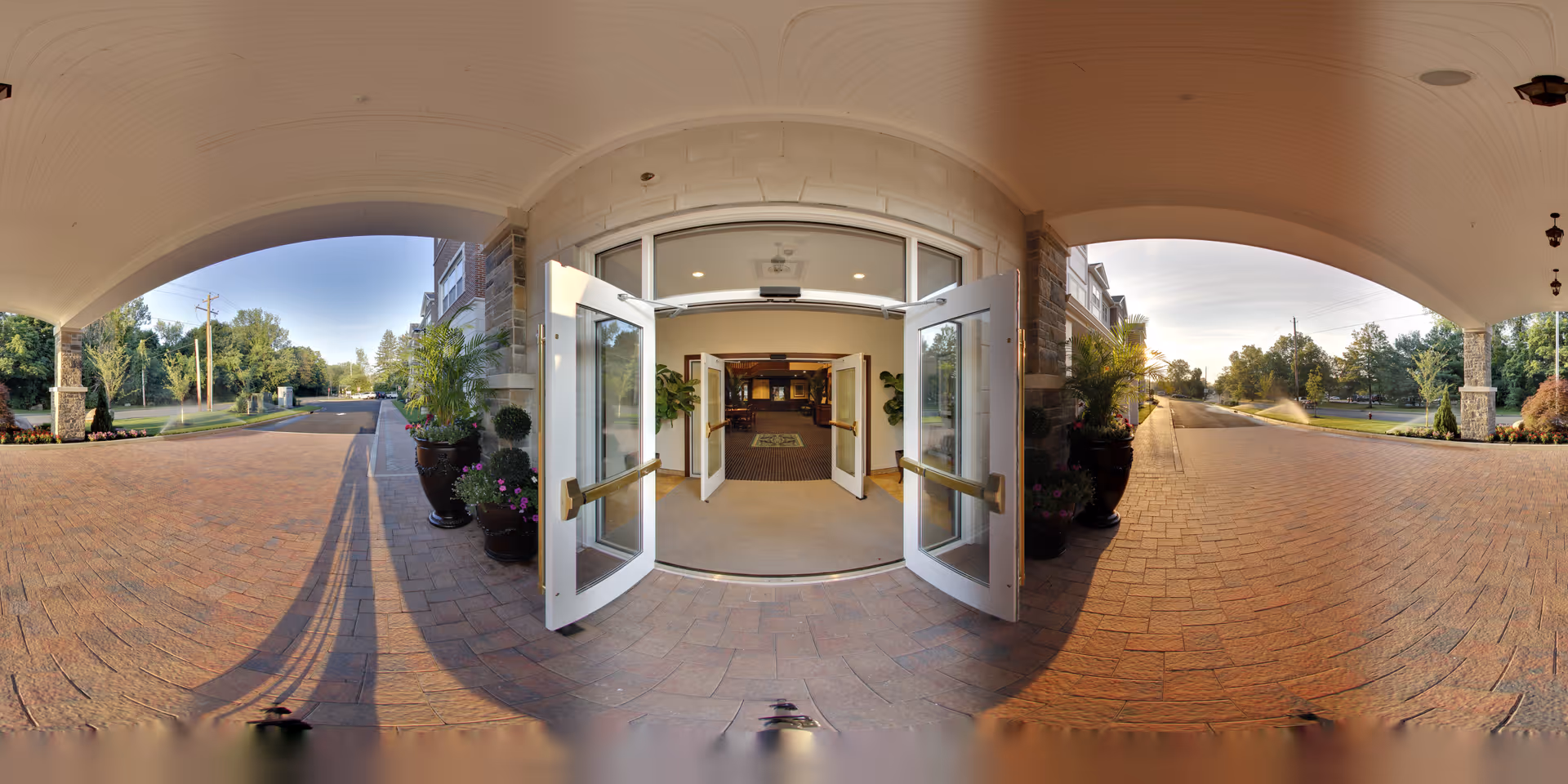 Wide-angle view of the entrance to a building with double glass doors open, showing a carpeted interior lobby area with plants on either side. The exterior features a covered brick-paved driveway with large planters and greenery on both sides, under a curved ceiling with recessed lighting. Trees and a road are visible in the background under a clear sky.