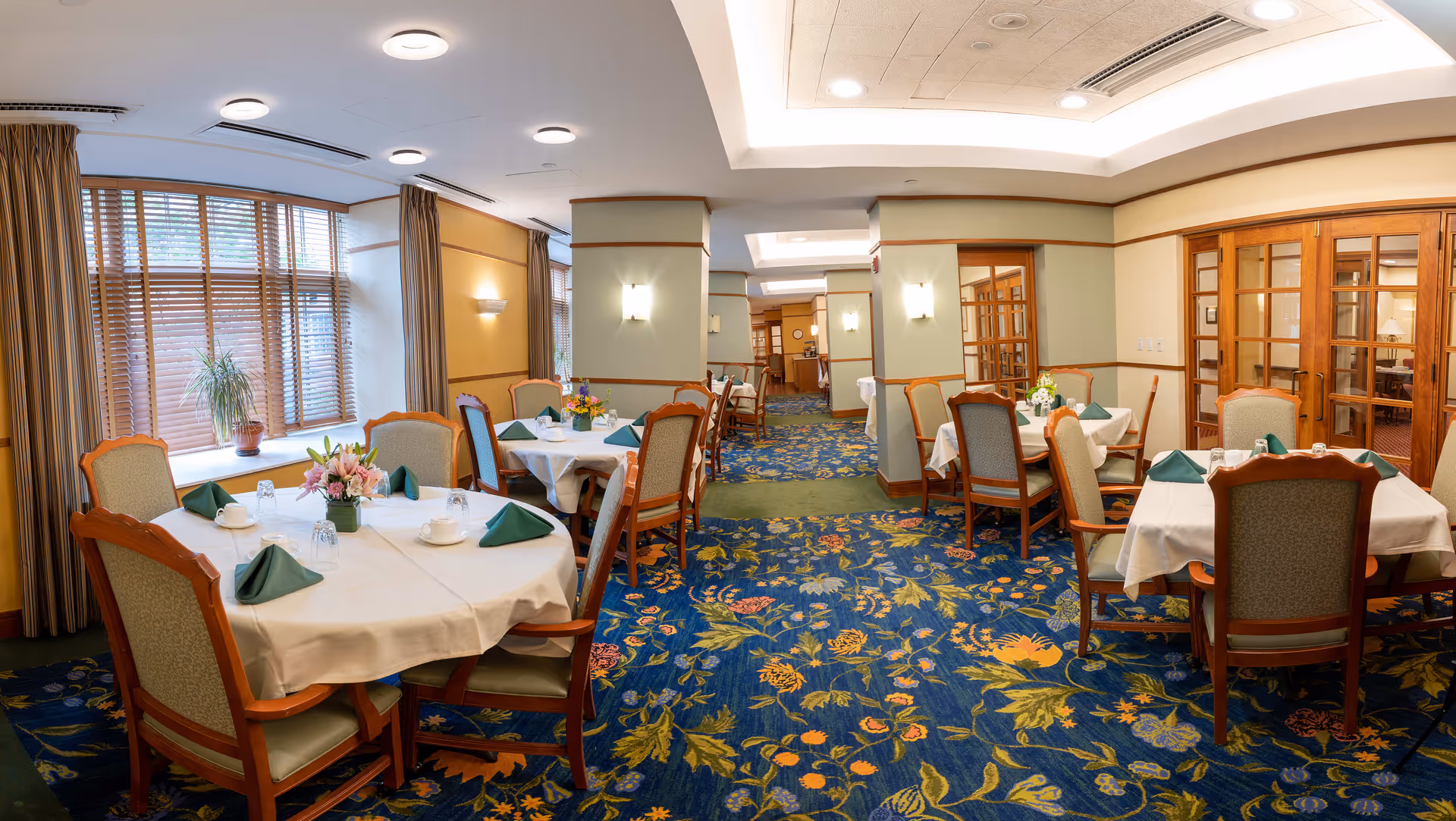 A well-lit dining room with multiple tables covered in white tablecloths, each set with green folded napkins, cups, and glasses. The room features large windows with wooden blinds and curtains, floral carpet, wooden chairs with cushioned seats, and soft wall lighting. There are flower arrangements on the tables and wooden framed glass doors on the right side.