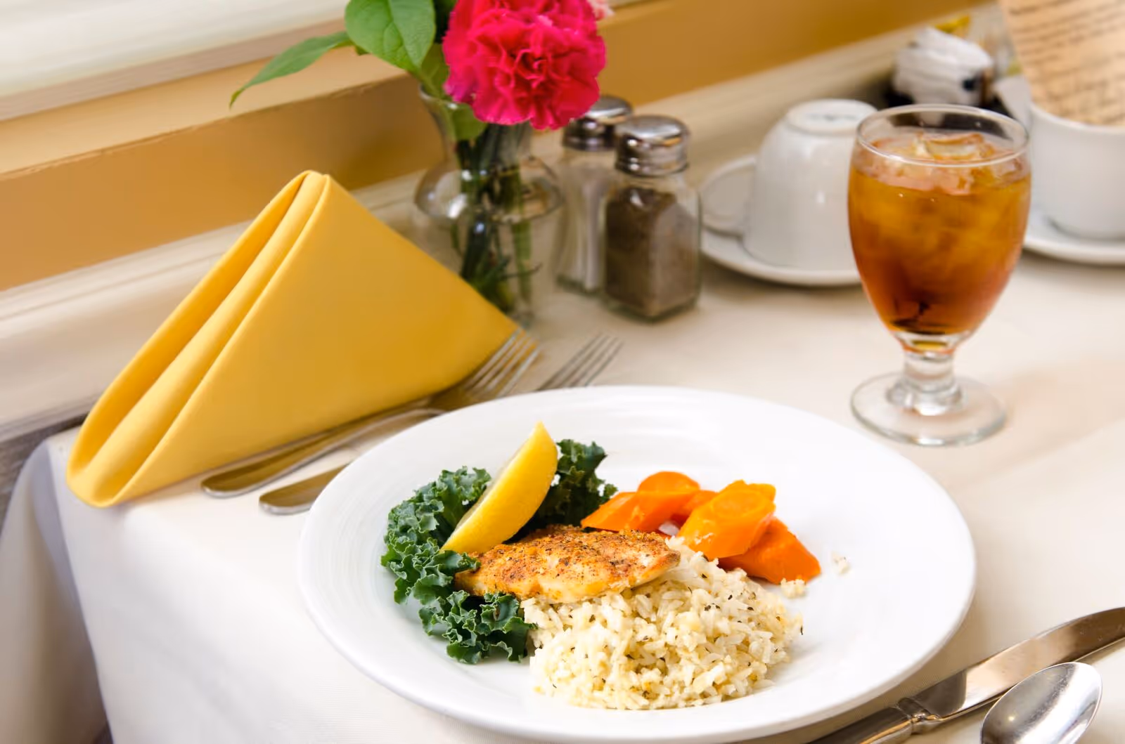 A neatly set dining table with a plate of food including seasoned rice, cooked carrots, a piece of grilled fish garnished with a lemon wedge and kale. There is a glass of iced tea, salt and pepper shakers, a yellow folded napkin, and a small vase with a pink flower on the table.