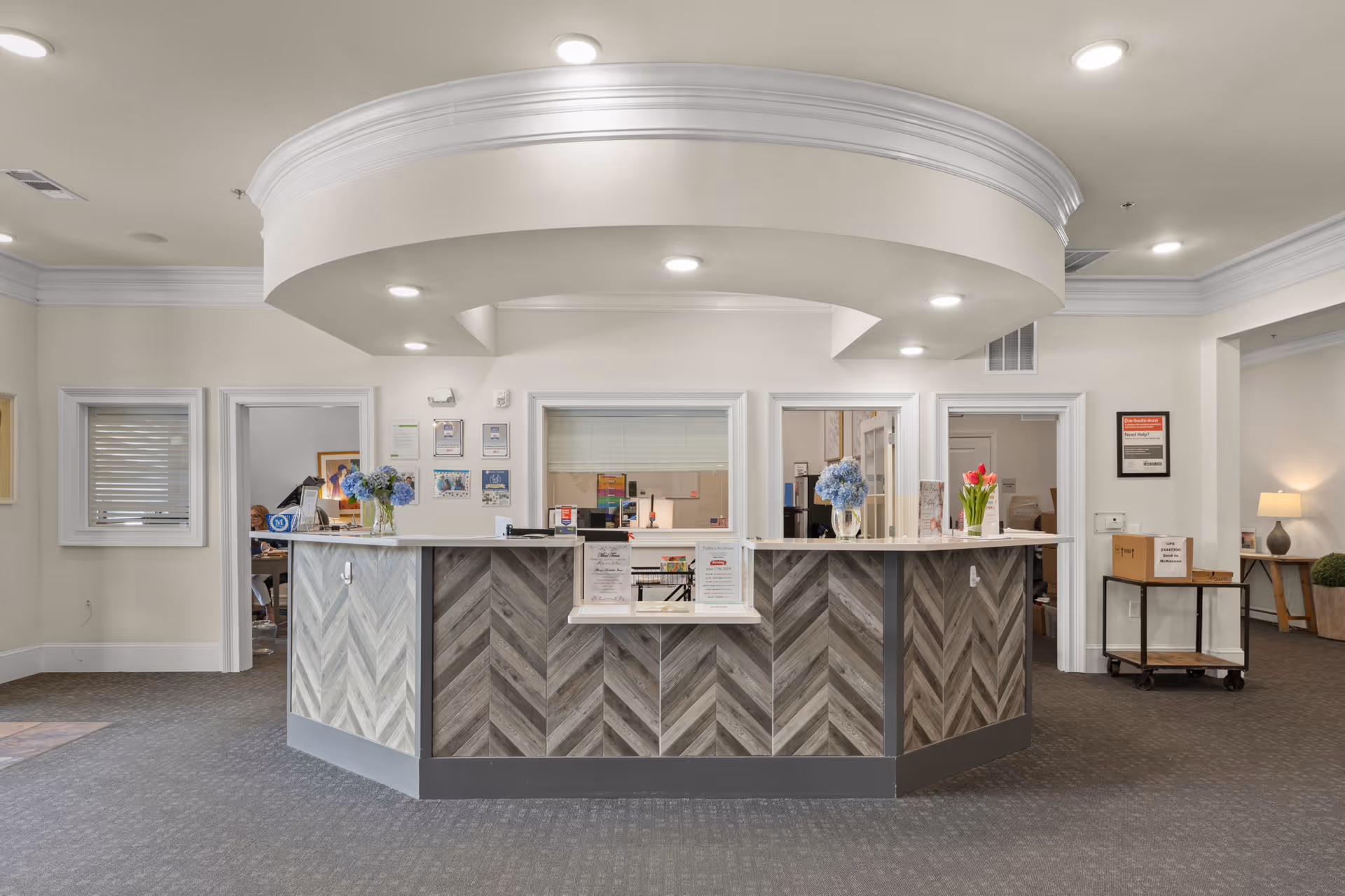 Reception desk area in a senior living facility with a curved ceiling design and recessed lighting. The desk features a chevron wood pattern and has vases with flowers on top. Behind the desk are windows and doorways leading to other rooms. The surrounding area has neutral-colored walls and carpeted flooring.