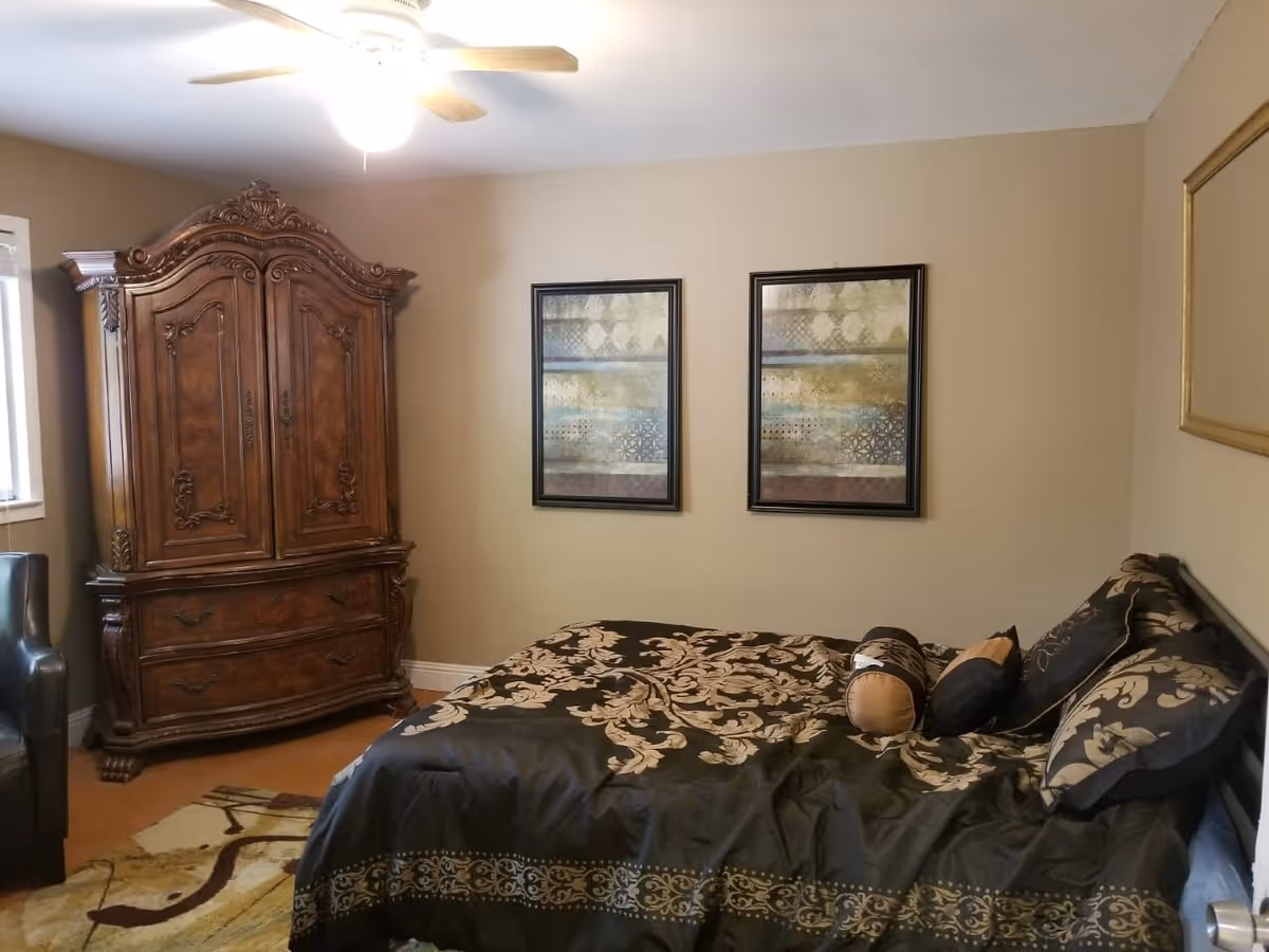 A bedroom with a large wooden armoire, a bed with black and gold patterned bedding and multiple pillows, two framed abstract paintings on the beige wall, a ceiling fan with light, a window, a black armchair, and a patterned area rug on the floor.
