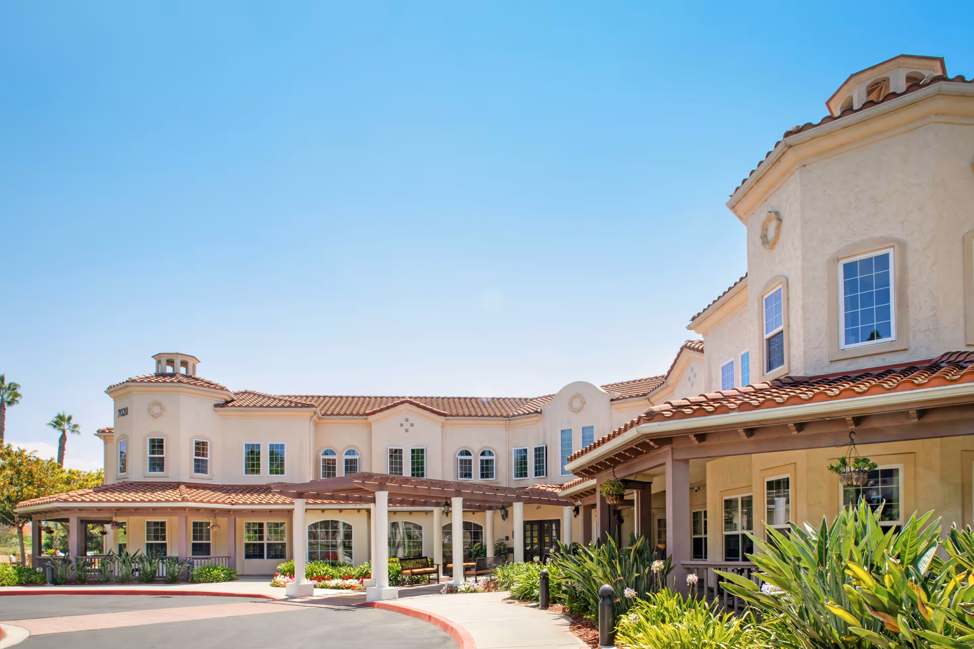 Exterior view of a large, two-story senior living facility with beige stucco walls and red tile roofing under a clear blue sky. The building features multiple windows, a covered entrance with columns, and well-maintained landscaping with green plants and flowers.