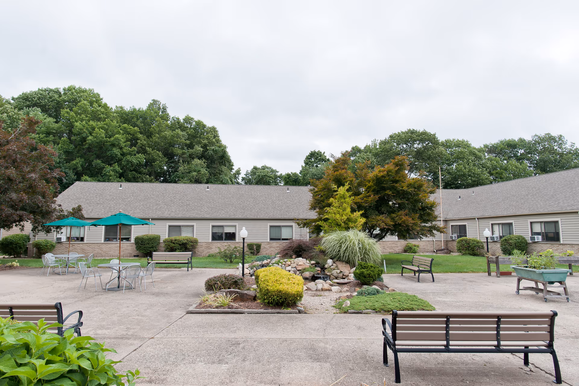 Courtyard with benches, patio tables with umbrellas, and a landscaped rock garden in front of a one-story residential building.
