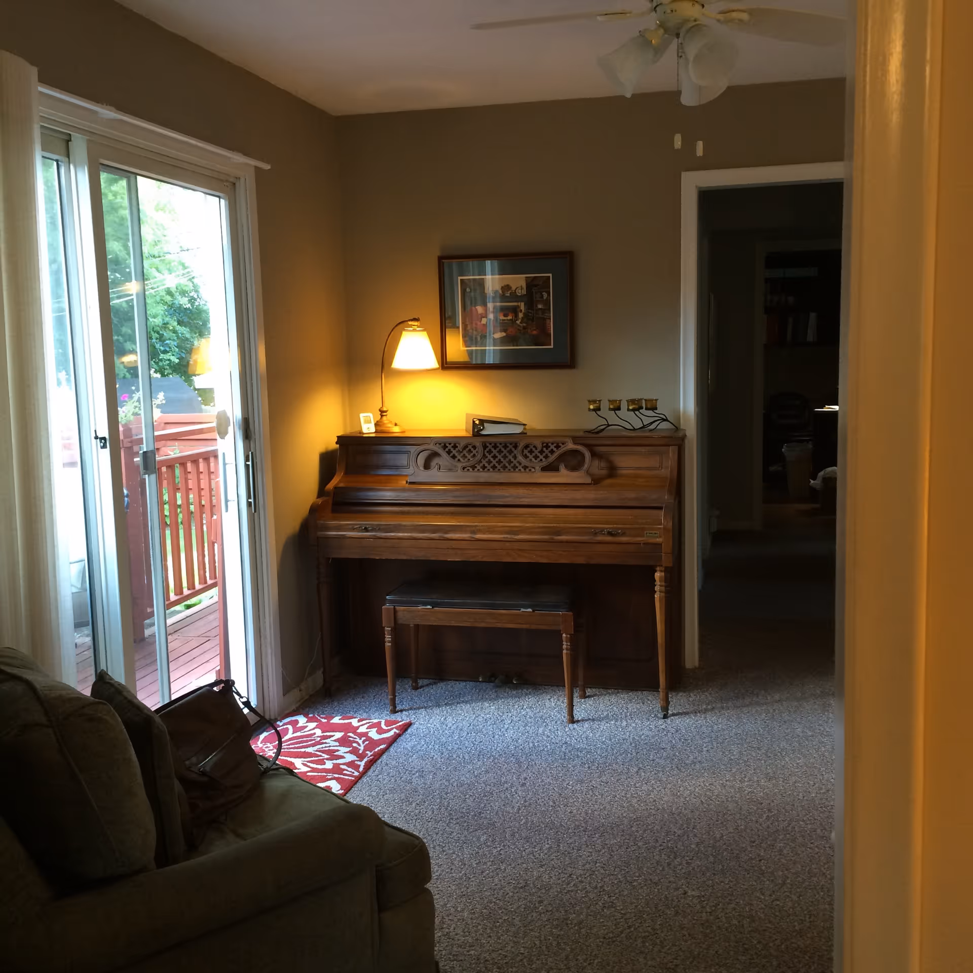 Interior view of a living room area with a wooden upright piano against a beige wall, a small lamp on top of the piano, a framed picture hanging above it, a sliding glass door leading to an outdoor deck, and a green couch partially visible on the left side.