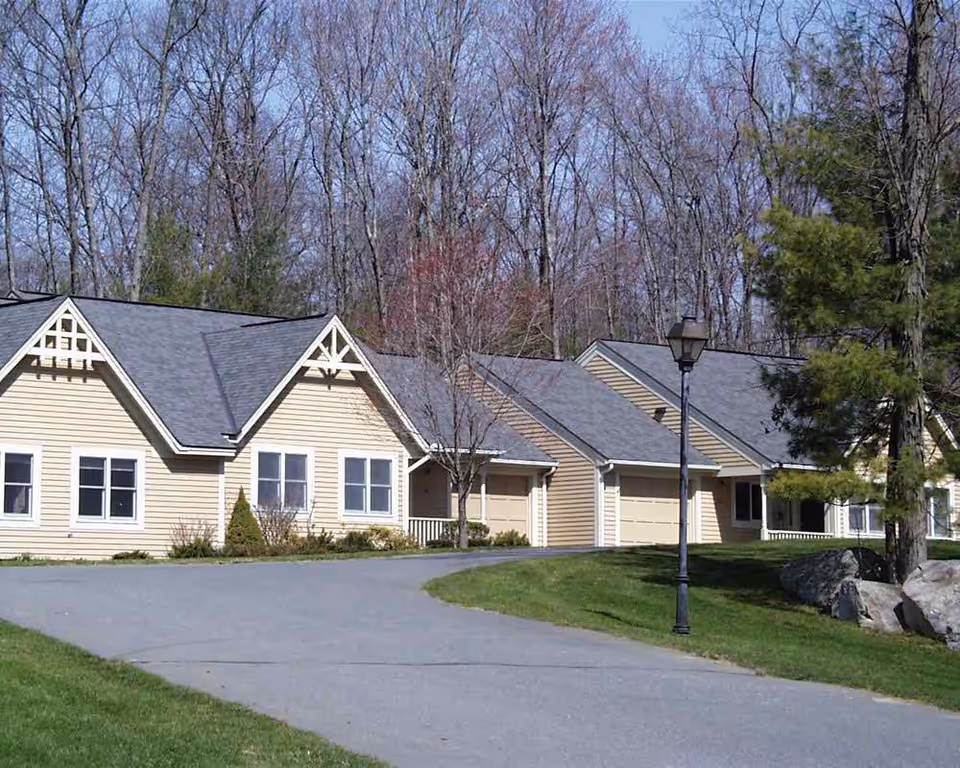 Row of single-story beige retirement community units with garages, a driveway, a lamp post, and trees in the background.