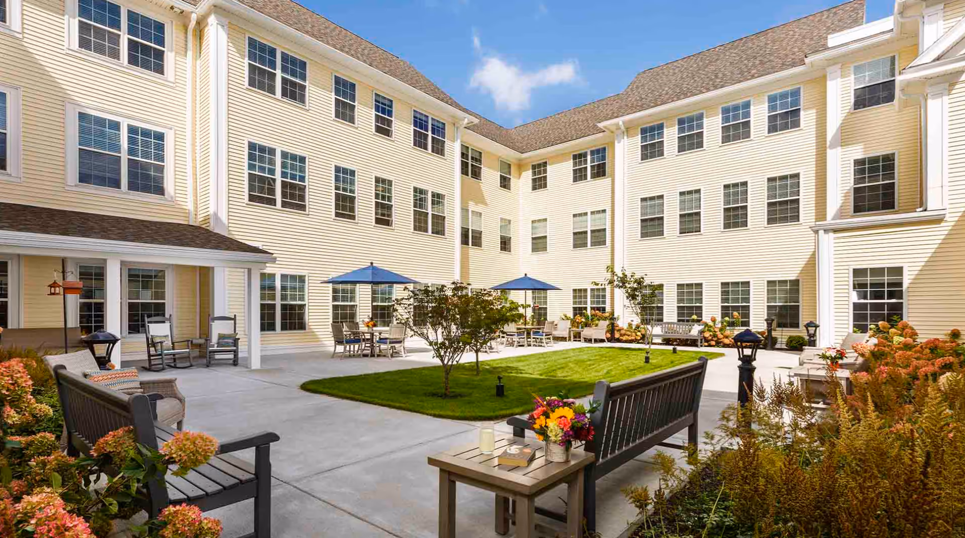 A bright outdoor courtyard area of a senior living facility with beige multi-story buildings surrounding a central green lawn. There are benches, tables with blue umbrellas, chairs, and various plants and flowers around the paved patio area under a clear blue sky.