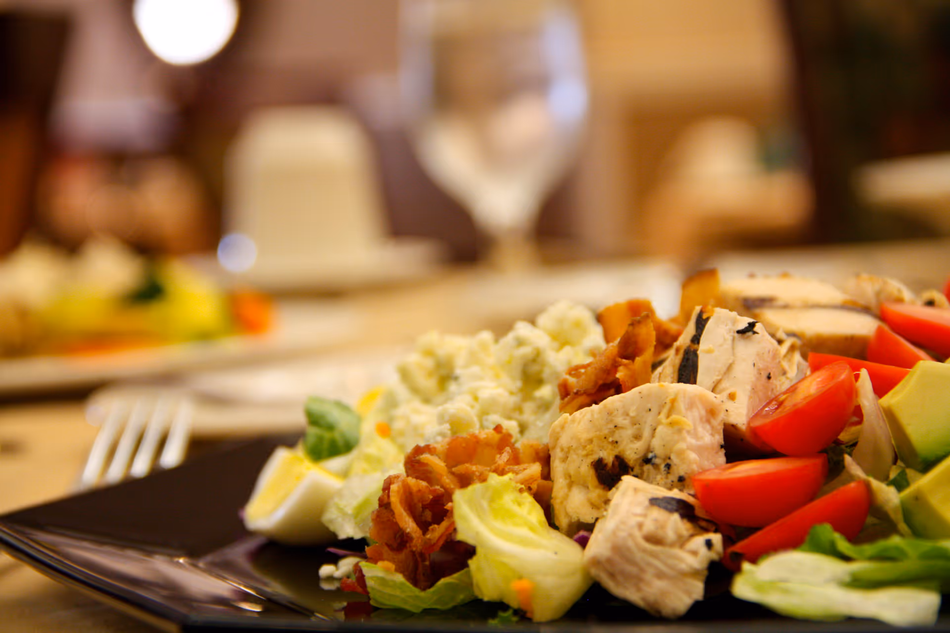 Close-up of a plate with a fresh salad including grilled chicken pieces, cherry tomatoes, avocado, bacon bits, lettuce, and a boiled egg, with a blurred background showing another plate and a glass of water.