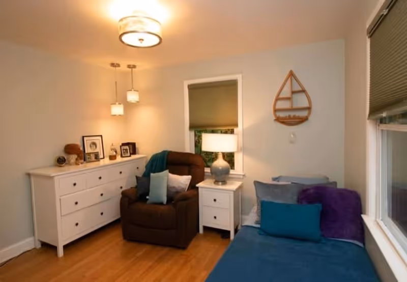 A cozy living room with a brown recliner chair adorned with multiple pillows, a white dresser with framed photos and decorative items, a white side table with a lamp, a blue daybed with pillows and a purple throw blanket, wooden flooring, and a window with a closed blind. A wooden sailboat wall decoration hangs on the wall.