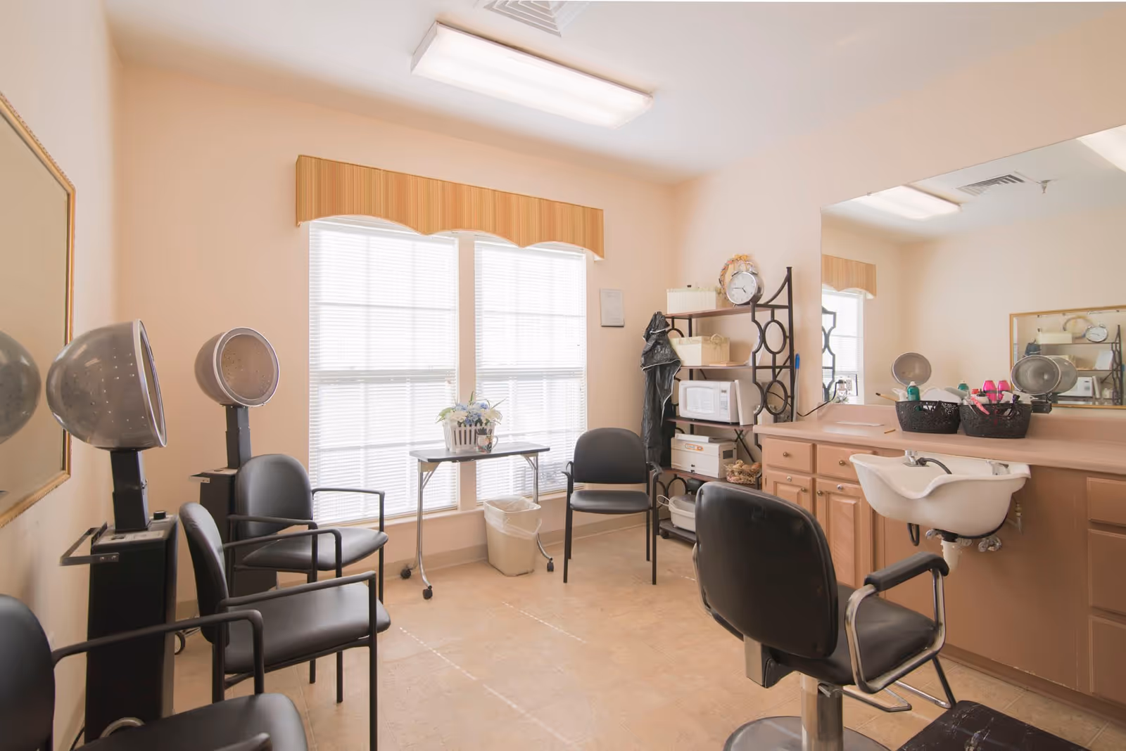 A small salon room with three black salon chairs, two hair dryers, a large mirror above a sink, a shelving unit with a microwave and baskets, and a window with blinds and a valance letting in natural light.