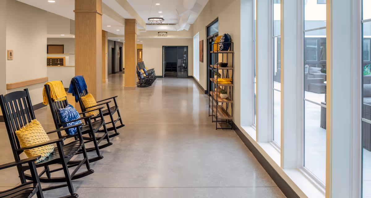 A bright hallway in a senior living facility with black rocking chairs lined up along the left wall, each adorned with colorful cushions and blankets. Large windows on the right side let in natural light and offer a view of an outdoor seating area. Wooden columns and a shelving unit with decorative items are also visible.