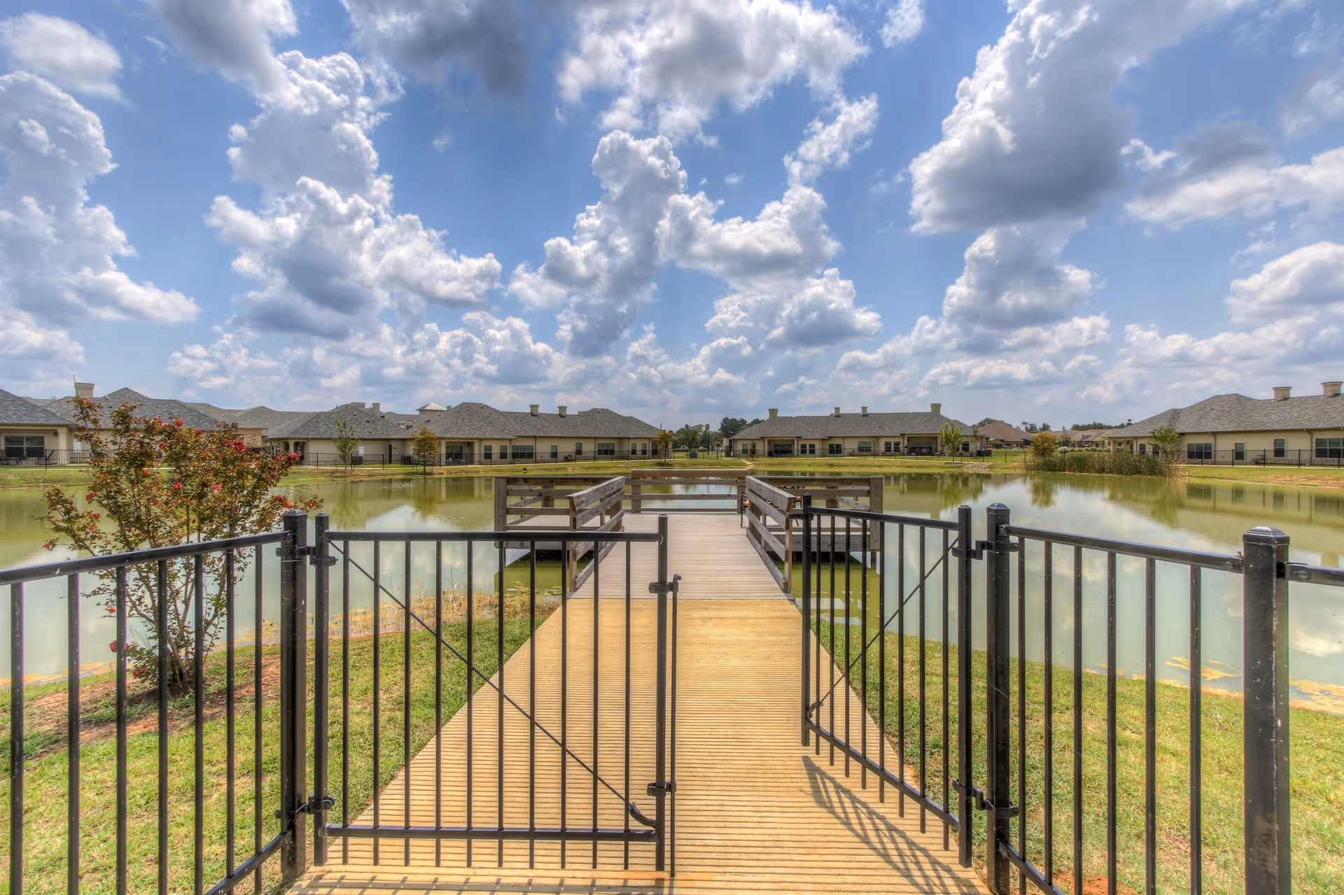 A wooden dock with railings extends over a calm pond, surrounded by a black metal fence with a gate in the foreground. Single-story buildings with gray roofs are visible around the pond under a partly cloudy blue sky.