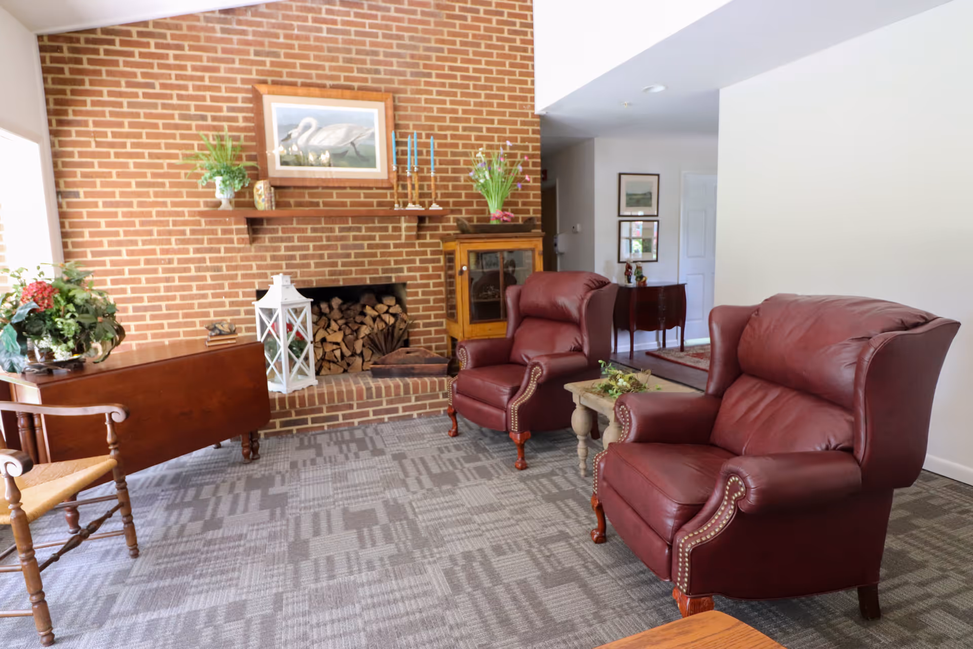 A cozy living room with two burgundy leather armchairs, a wooden side table with a floral arrangement, a brick fireplace filled with stacked firewood, and a wooden cabinet. The room has a patterned carpet and light-colored walls, with framed artwork and plants decorating the space.