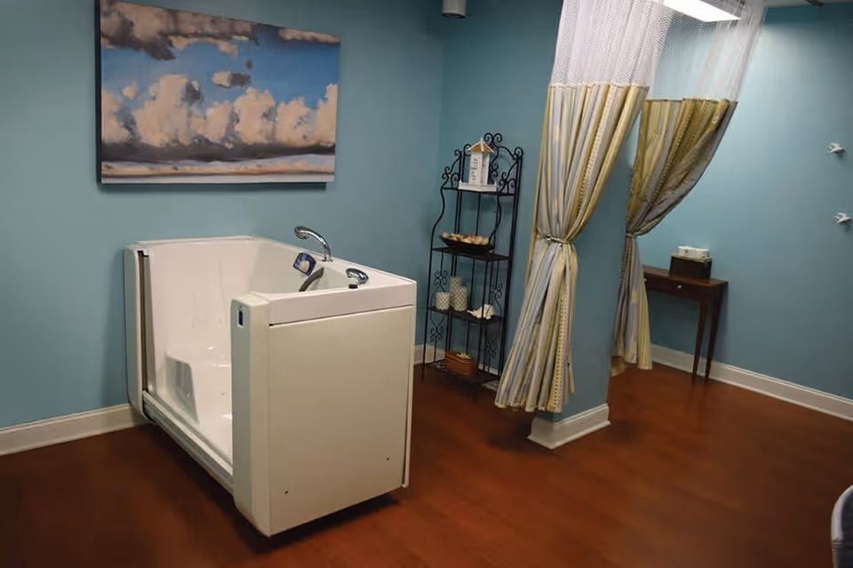 A room with light blue walls and wooden flooring featuring a white walk-in bathtub with a faucet. There is a painting of clouds on the wall above the bathtub, a black metal shelf with decorative items, and a small wooden table with a tissue box. Two striped curtains are tied back, partially covering a doorway.