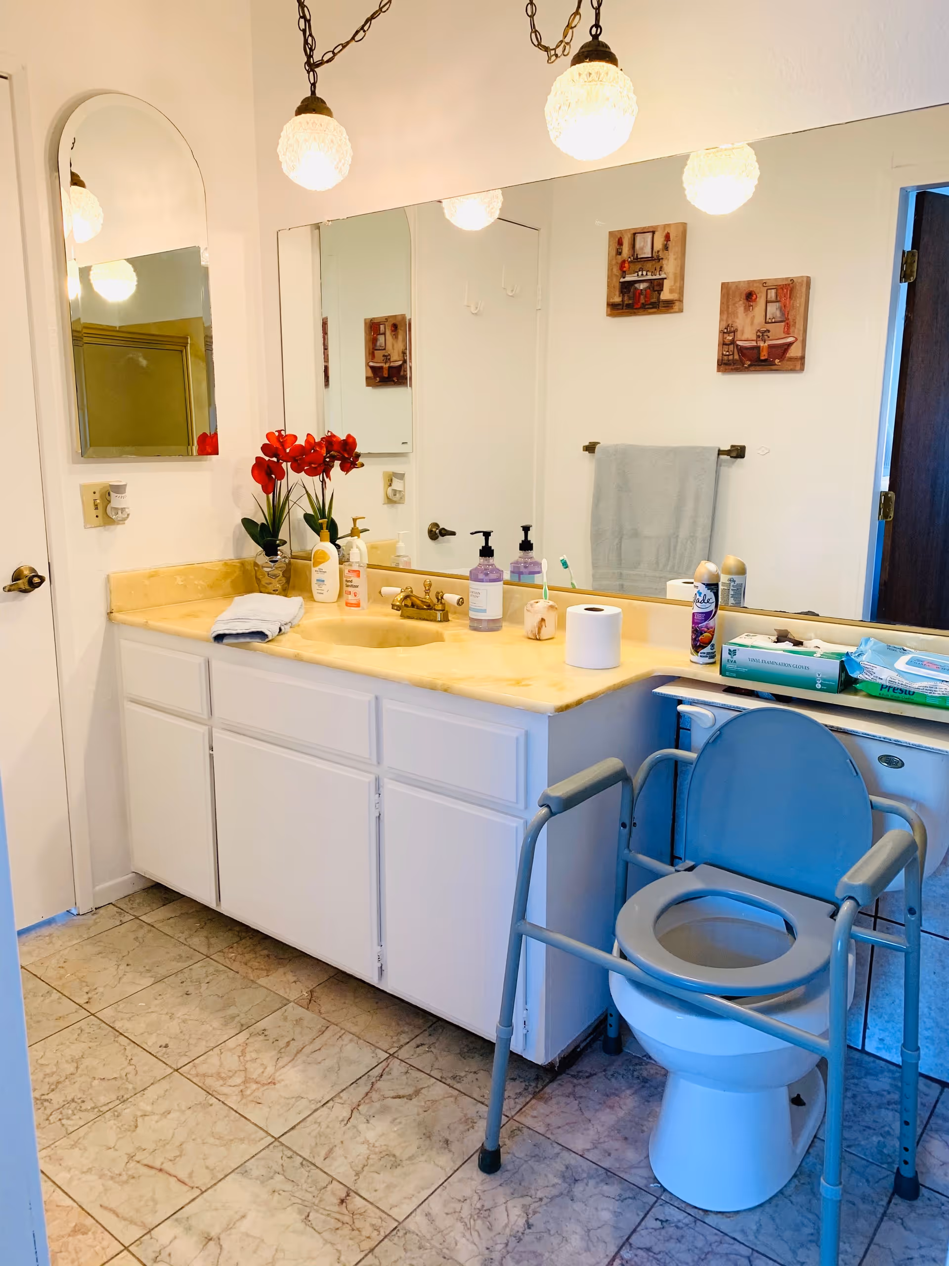 A bathroom with a large mirror above a yellow countertop with a sink. On the countertop are various toiletries, a roll of toilet paper, and a vase with red flowers. There is a blue raised toilet seat with armrests positioned over the toilet. The floor is tiled, and two framed pictures hang on the wall above a towel rack with a light blue towel.