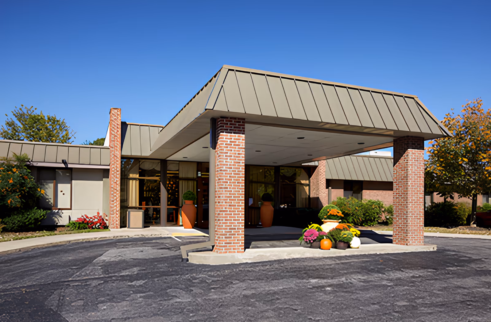 Entrance of a senior living facility with a covered drop-off area supported by brick pillars. There are large planters with greenery and colorful flowers near the entrance. The building has a beige and brick exterior with a clear blue sky above.