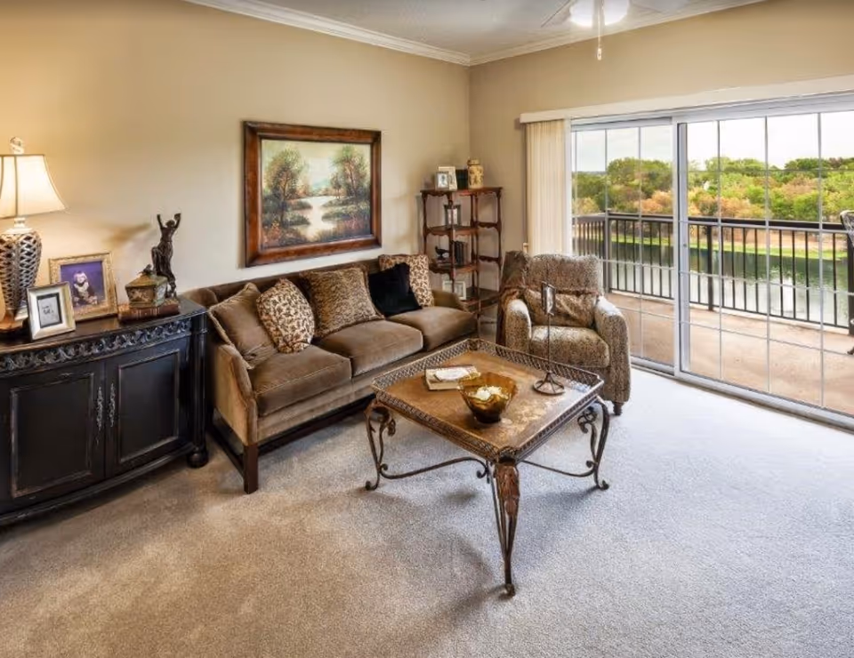 A cozy living room with a brown sofa adorned with patterned cushions, a matching armchair, and a decorative coffee table with items on it. There is a black sideboard with a lamp, framed photos, and a small statue. A large framed painting of a nature scene hangs on the beige wall. Sliding glass doors open to a balcony with a view of trees and a body of water outside.