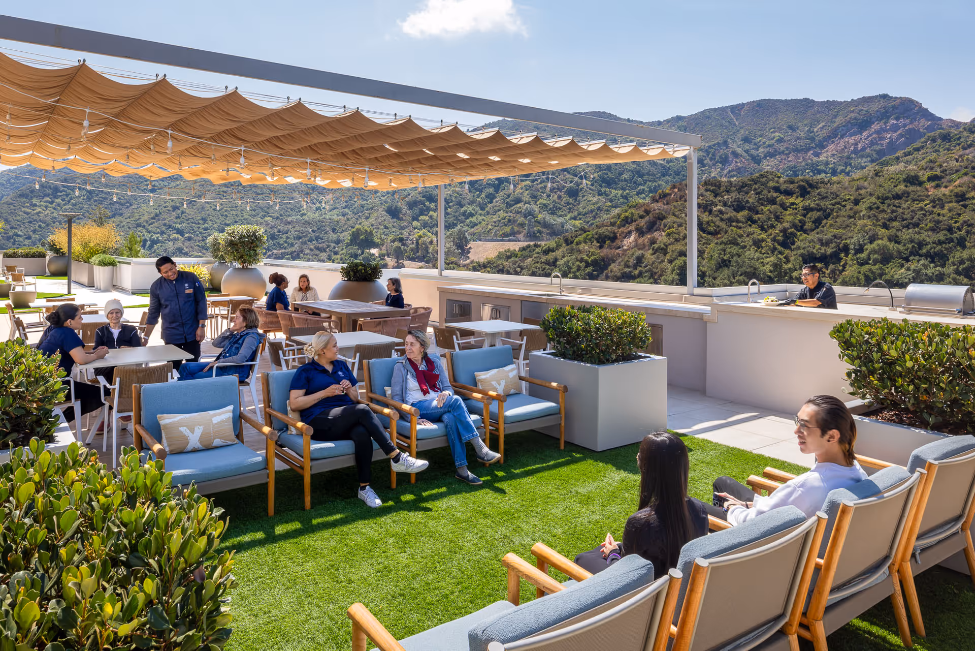 A group of people sitting and chatting on a rooftop patio with comfortable chairs and tables under a fabric canopy. The area is decorated with large potted plants and overlooks a scenic mountainous landscape under a clear blue sky.
