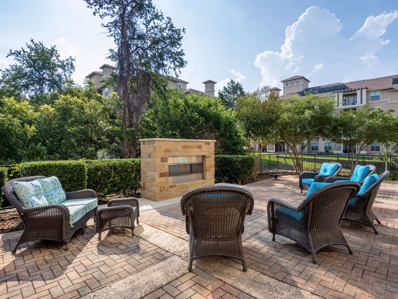 Outdoor patio area with wicker chairs and sofas arranged around a stone fireplace, surrounded by greenery and trees, with a multi-story building in the background under a partly cloudy sky.