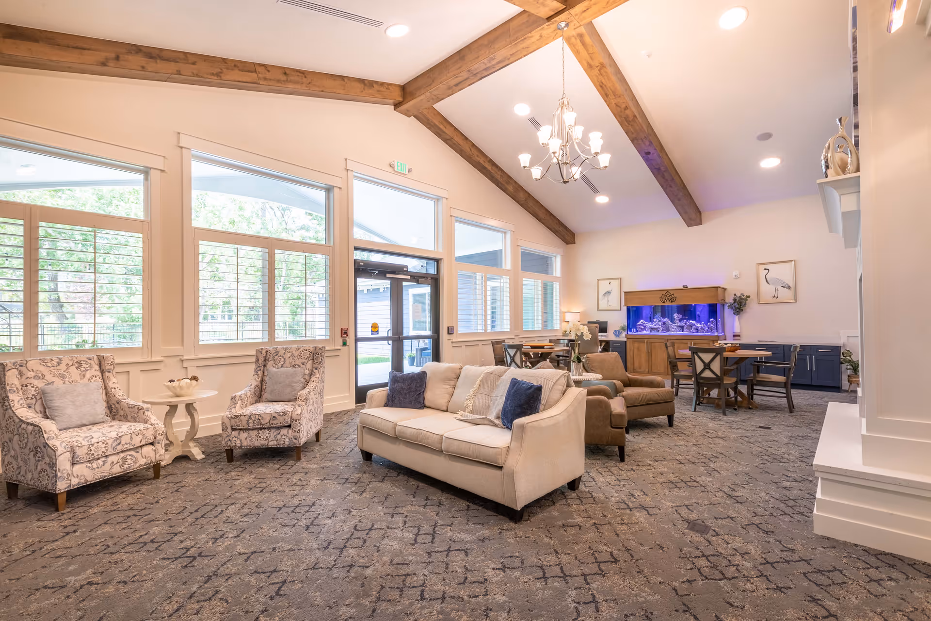 A spacious and well-lit senior living common area with high vaulted ceilings featuring wooden beams. The room includes a beige sofa with cushions, two patterned armchairs with a small round table between them, and two brown armchairs. There is a large fish tank against the far wall, flanked by framed bird artwork. Several tables and chairs are arranged near the fish tank, and large windows with white trim allow natural light to fill the room. The carpet has a patterned design, and a chandelier hangs from the ceiling.