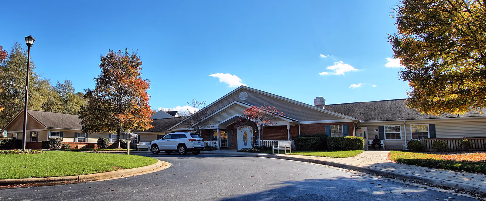 Exterior view of a single-story senior living facility building with a driveway in front, a white SUV parked near the entrance, surrounded by green lawns and trees with autumn foliage under a clear blue sky.