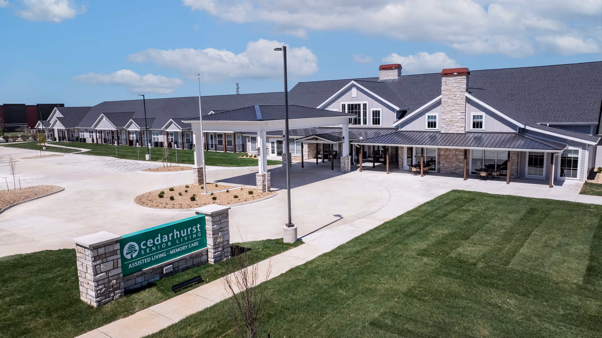 Exterior view of Cedarhurst Senior Living of Lawrence, showing a large building with a covered entrance, multiple windows, and a well-maintained lawn. A green sign in front reads 'Cedarhurst Senior Living Assisted Living - Memory Care'. The sky is clear with a few clouds.