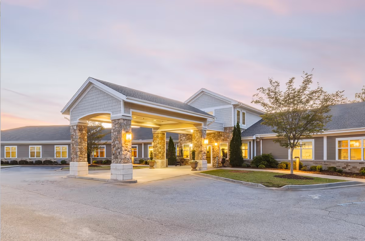 Exterior view of a senior living facility named Patewood Post-Acute at dusk, showing a covered entrance with stone pillars, well-lit windows, a tree, and a paved driveway.