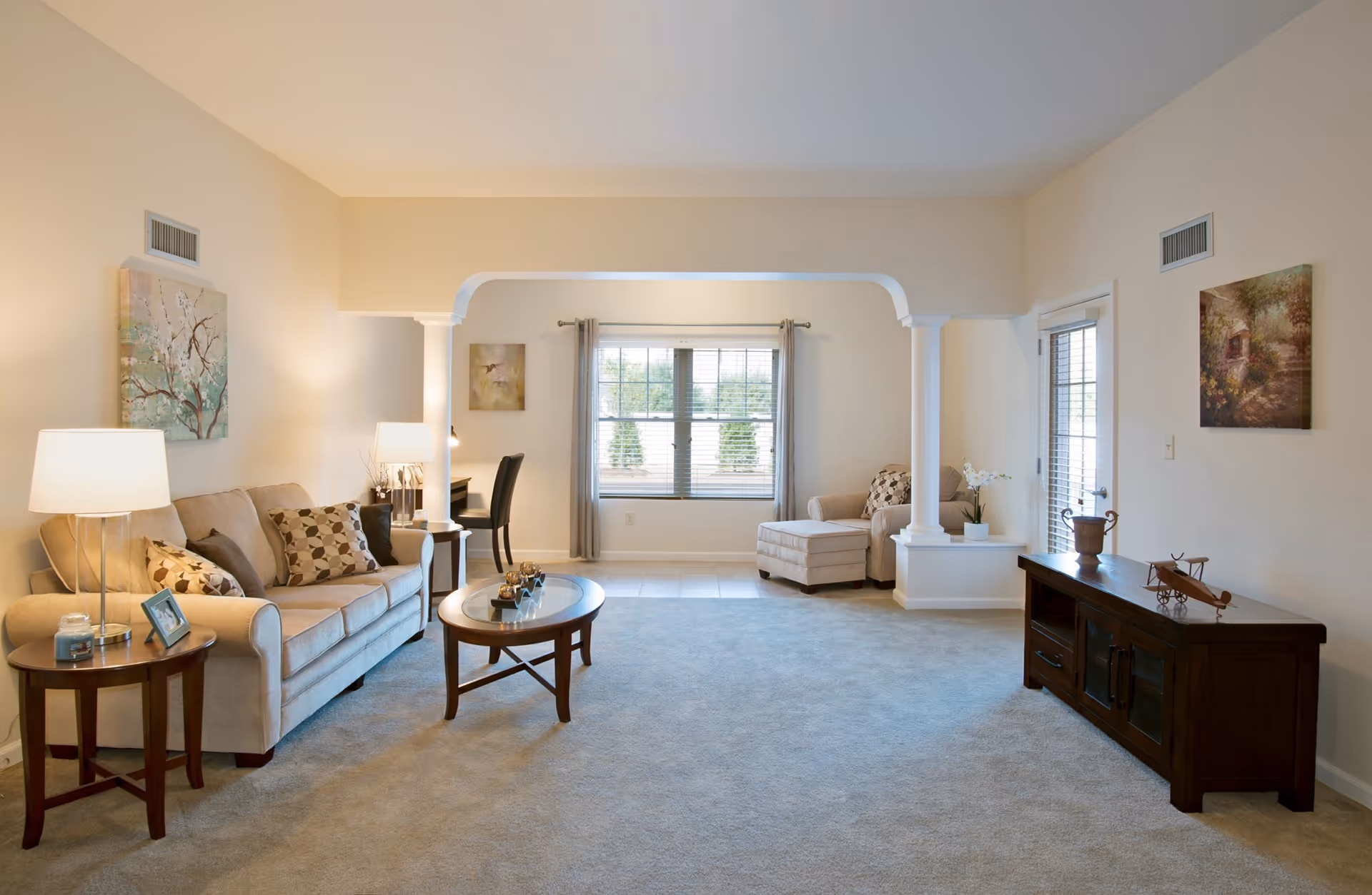 A spacious living room with beige walls and carpet. There is a beige sofa with patterned cushions on the left, accompanied by a wooden side table with a lamp and a photo frame. In front of the sofa is a wooden coffee table with decorative items. The room features two white columns framing a large window with gray curtains. To the right, there is a beige armchair with a matching ottoman and a wooden media console with decorative items on top. The room is well-lit with natural light coming through the window and a glass door on the right.