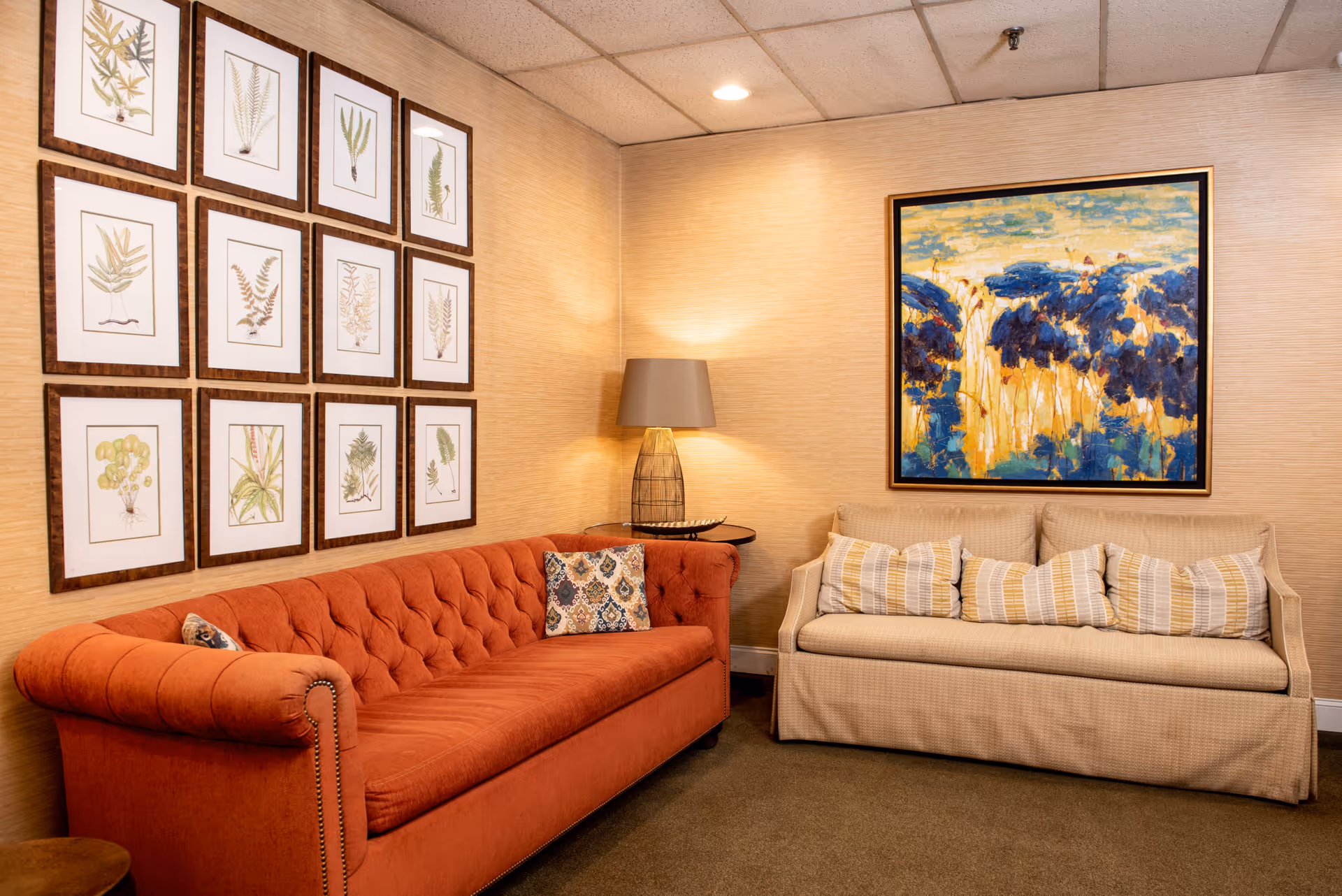 A cozy living room area with an orange tufted sofa and a beige sofa adorned with striped pillows. A table lamp with a beige shade sits on a small round table between the sofas. The walls are decorated with a grid of framed botanical prints on one side and a large colorful abstract painting on the other.