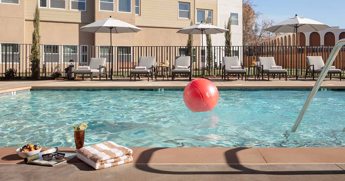 Poolside scene with a red ball floating in the water, towels and a drink on the deck, and lounge chairs with umbrellas in front of a building.