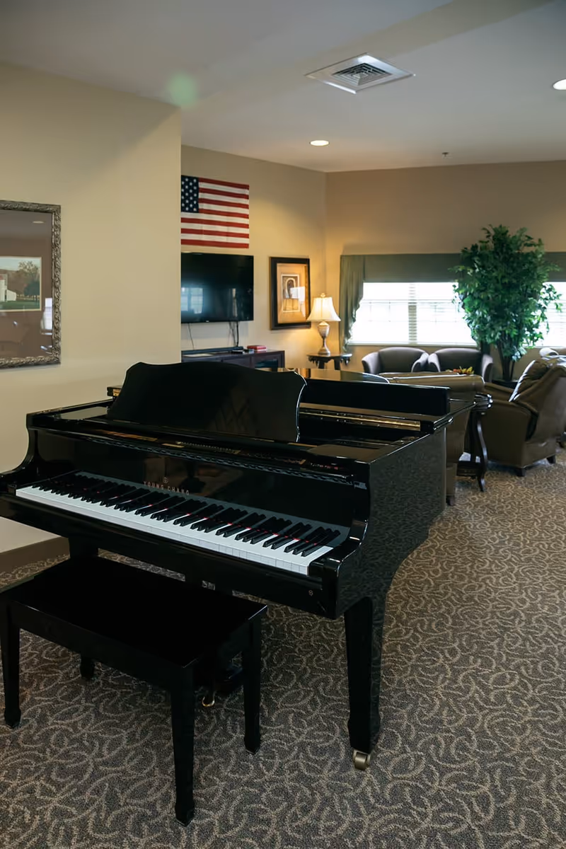 Interior of a senior living facility common area featuring a black grand piano with a matching bench on patterned carpet. In the background, there is a seating area with armchairs, a side table with a lamp, a large window with green curtains, a potted plant, a wall-mounted TV, an American flag, and framed artwork on beige walls.
