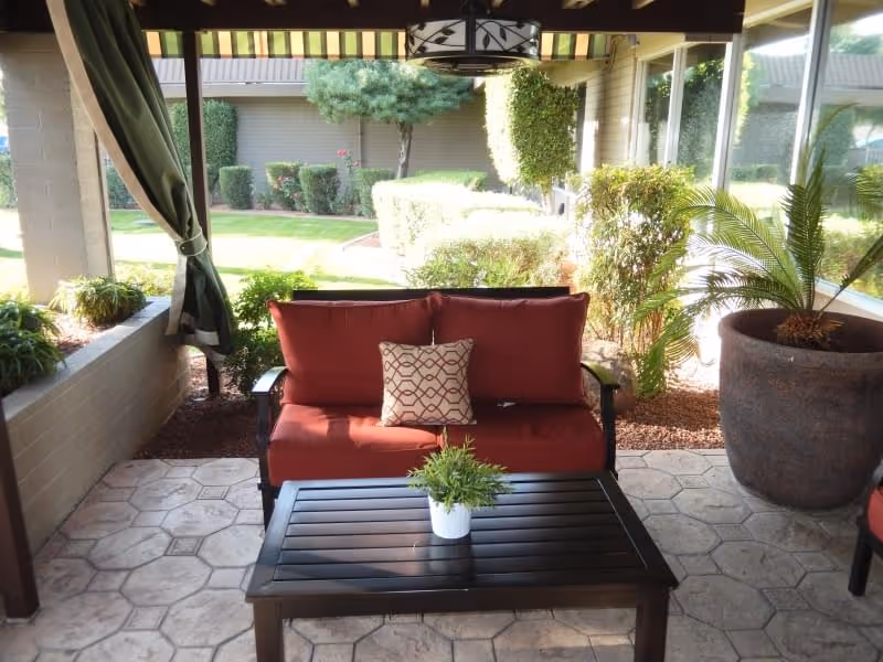 Outdoor covered patio area with a red cushioned loveseat, a decorative pillow, a black coffee table with a small potted plant, and large potted plants on the side. The patio overlooks a well-maintained garden with trimmed bushes and trees.